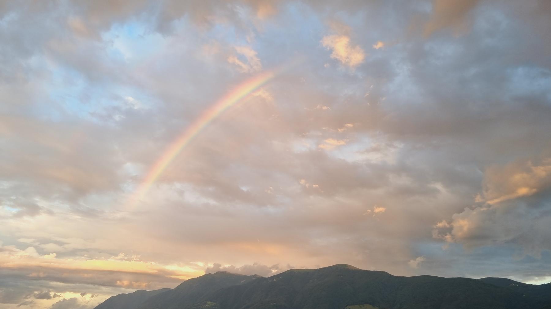 Zu sehen ist ein Regenbogen sder sich uber den Lago Maggiore spannt, er reflektiert im stillen, blau-grünen Wasser. Die Häuser von Brissago. Die Aussicht vom Casa Ramerino erstreckt sich fast über den ganzen Lago Maggiore.